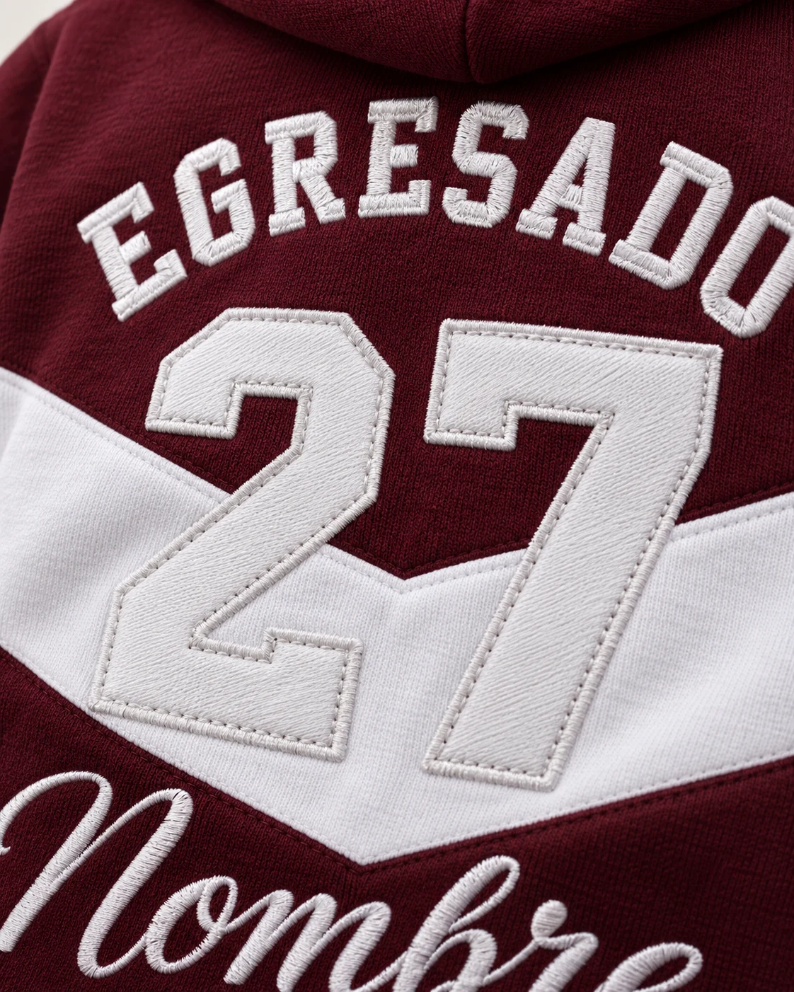 Campera bordó con franja blanca de frisa premium, letras bordadas y parche del año 27 cosido. Vista de detalle.